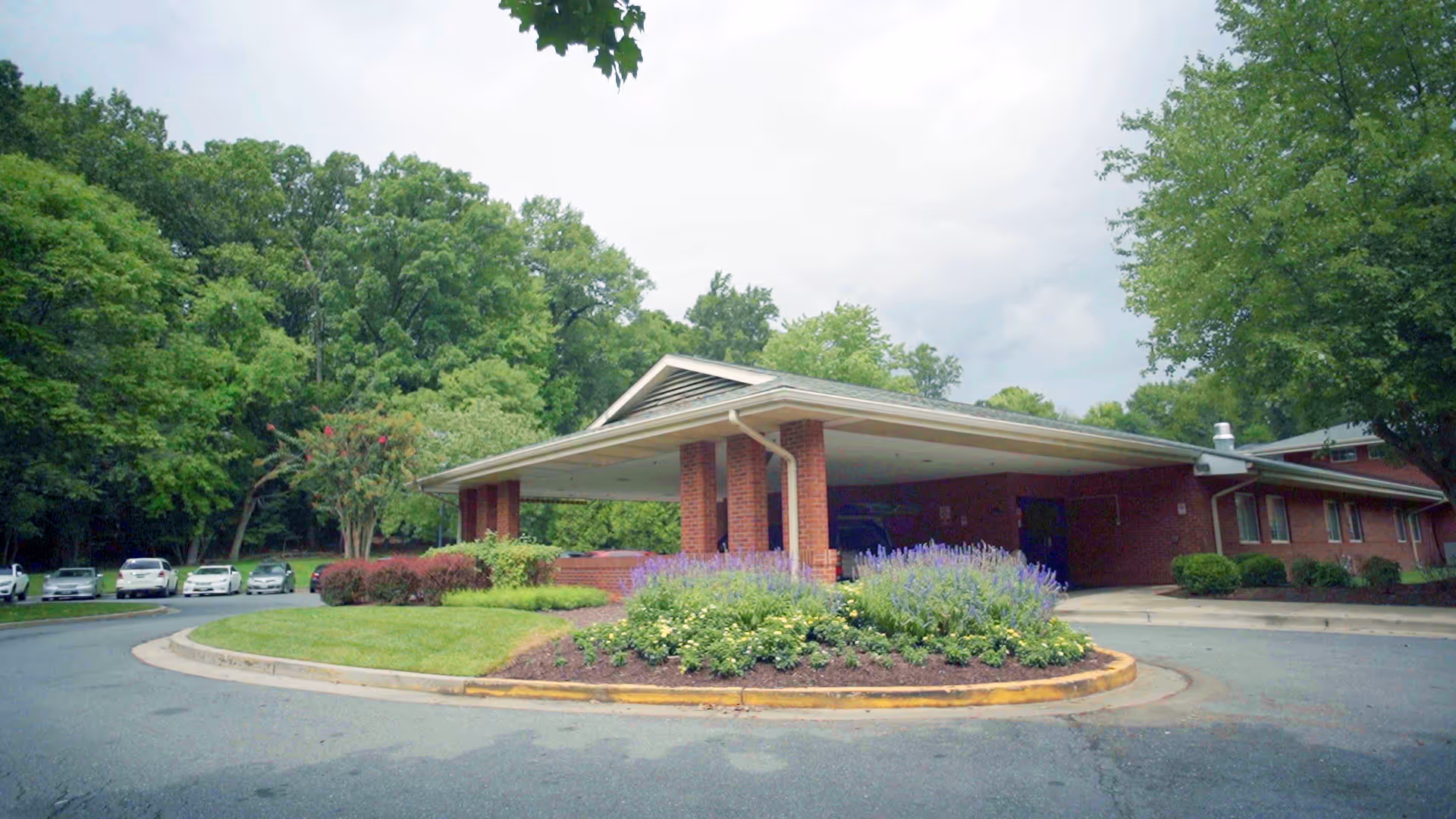 Front exterior view of Layhill Nursing And Rehabilitation Center showing a brick building with a covered entrance, surrounded by landscaped greenery and trees, with several parked cars visible in the background.
