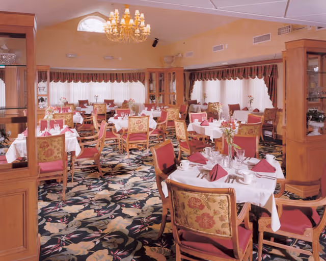 A formal dining room with multiple tables set with white tablecloths and burgundy napkins, floral-upholstered chairs, a chandelier, and patterned carpet.