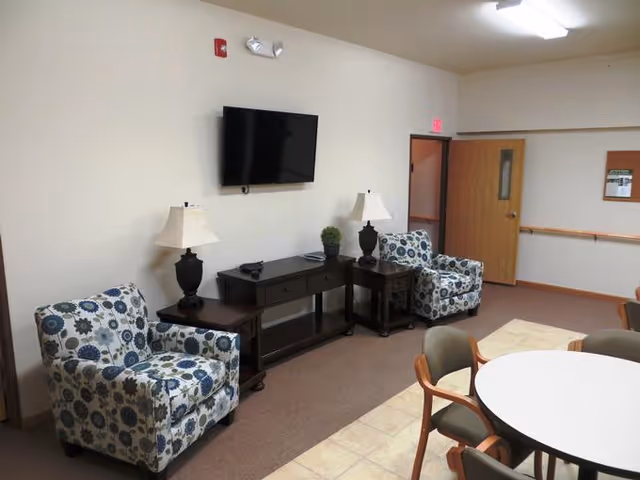 A common area in an assisted living facility featuring two patterned armchairs with floral designs, two side tables each with a lamp, a TV mounted on the wall above a console table, and a round table with chairs in the foreground. The room has beige walls, a tiled floor near the table, and carpet near the seating area. An open door and a bulletin board are visible in the background.