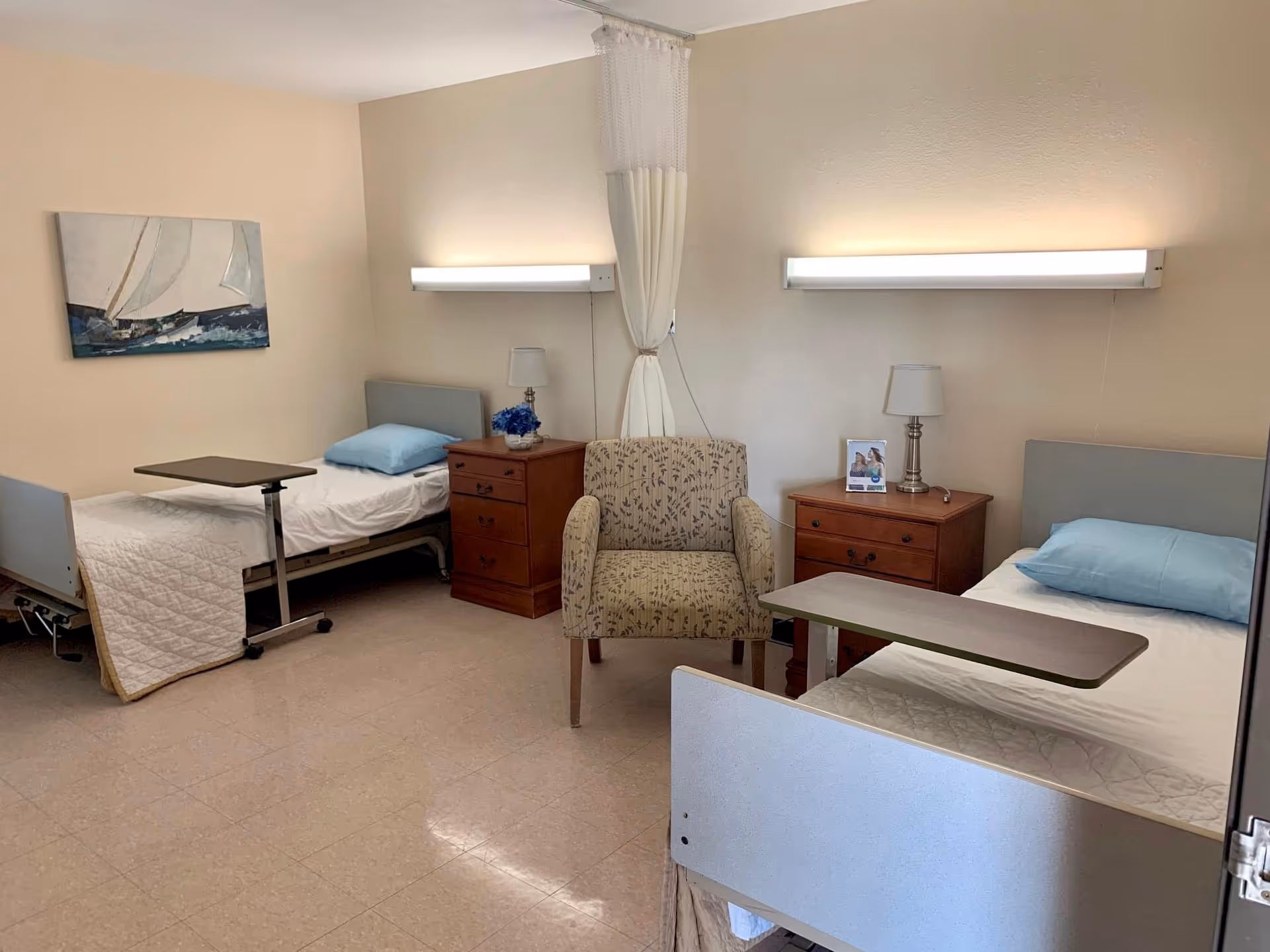 A nursing facility room with two hospital beds, each with a blue pillow and white bedding. Between the beds are two wooden nightstands with lamps and a small framed photo. A patterned armchair is placed between the beds. A painting of a sailboat hangs on the wall above one bed. The room has beige walls and tiled flooring.