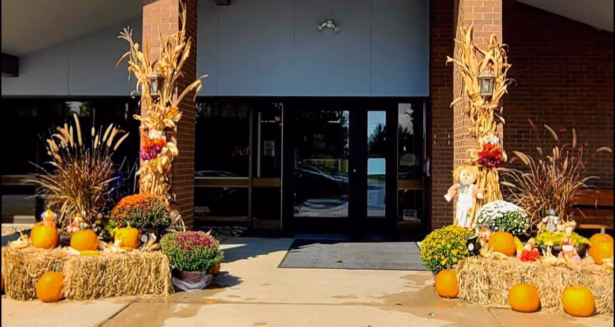 Entrance of a building decorated with autumn-themed items including pumpkins, hay bales, colorful flowers, corn stalks, and a scarecrow doll on either side of the doorway.