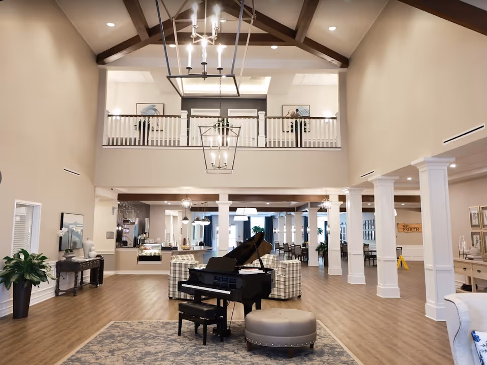 Two-story community lounge with a grand piano, seating areas, columns and a vaulted ceiling with chandeliers.