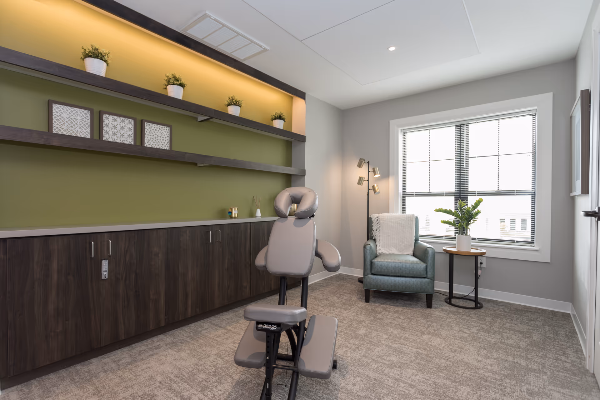 A well-lit room with a massage chair in the center, a comfortable armchair with a white throw blanket near a window, a small round side table with a potted plant, and dark wooden cabinets with shelves above holding small potted plants and framed art pieces on a green accent wall.