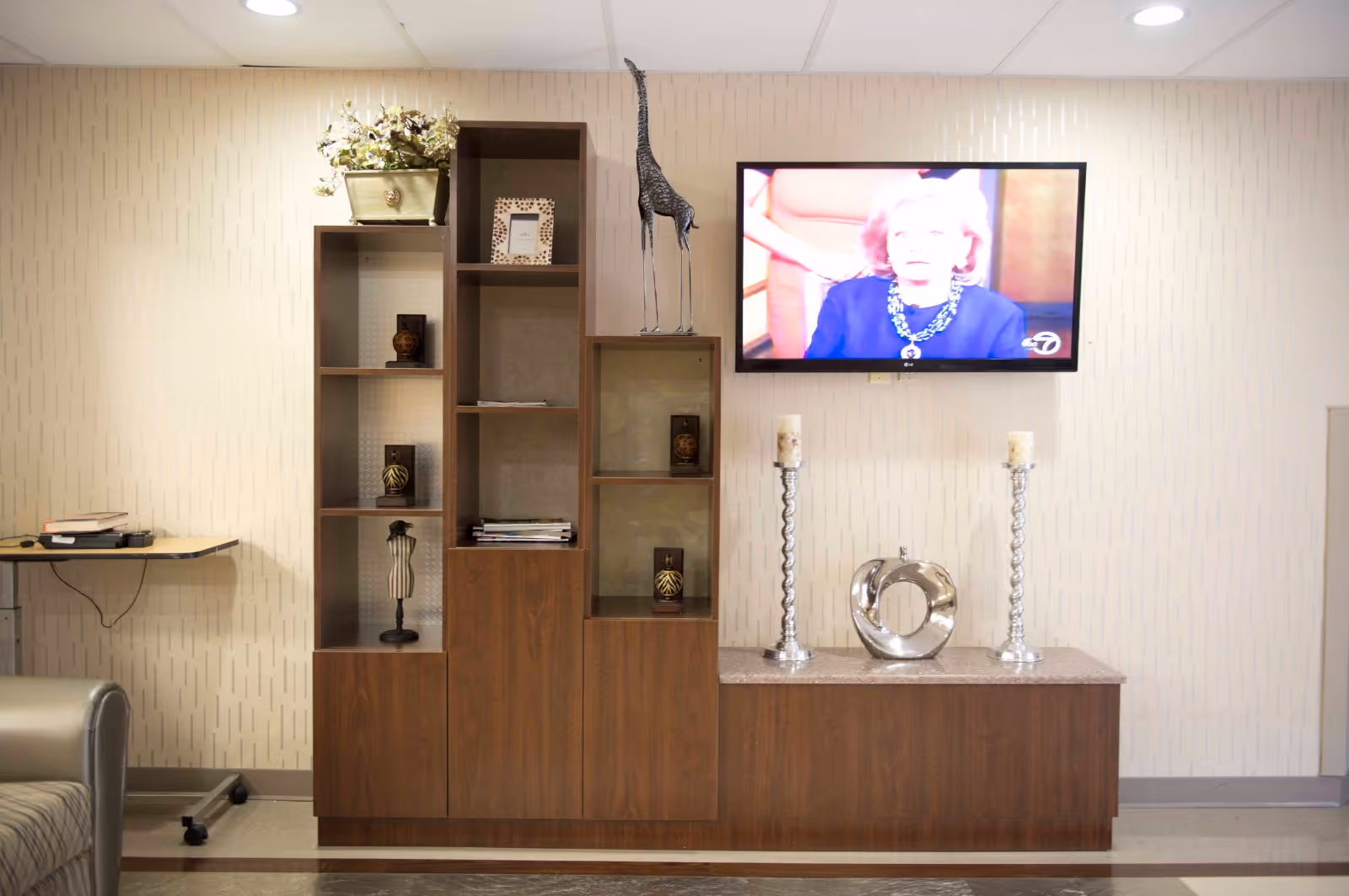 A living room area with a wooden shelving unit and cabinet against a beige patterned wall. The shelves hold decorative items including a potted plant, picture frame, small sculptures, and candles. A flat-screen TV mounted on the wall displays a woman speaking. Two tall silver candle holders and a silver decorative sculpture are placed on the cabinet below the TV. Part of a gray armchair and a small table with books and a phone are visible on the left side.
