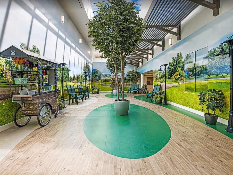 Indoor seating area with green benches and potted trees, surrounded by walls decorated with large murals of a park with green grass and trees. The floor has a wood-like pattern with green oval shapes, and there is a small cart with flowers and baskets on the left side. The ceiling has skylights and metal beams.