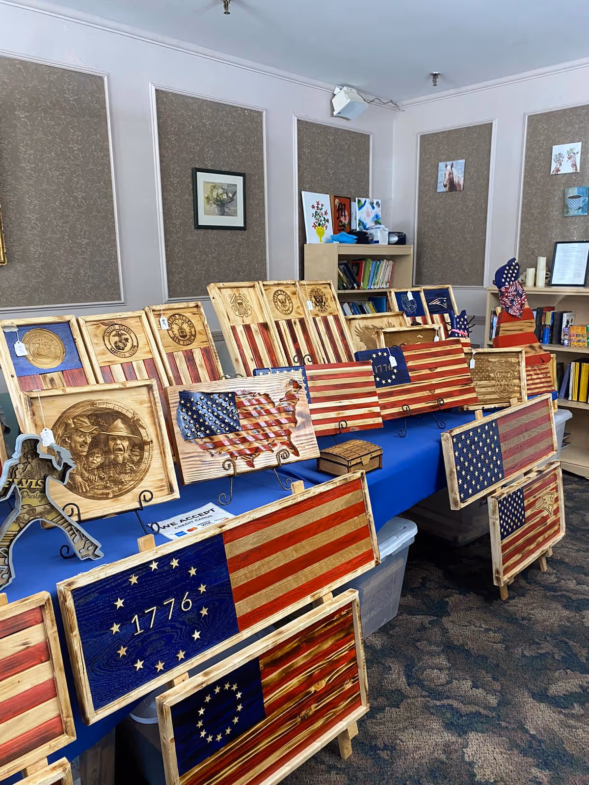 A display table covered with a blue cloth showcasing various wooden plaques and signs featuring American flag designs, patriotic symbols, and military emblems. The background includes bookshelves with books and framed pictures on the walls in a room with carpeted flooring.