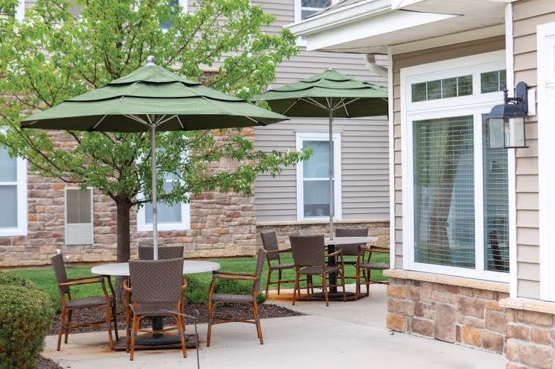 Outdoor patio area with two round tables, each shaded by a green umbrella. Each table is surrounded by four brown wicker chairs. The patio is adjacent to a building with beige siding and stone accents, and there is a tree and some bushes nearby.