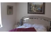 A bedroom with a bed featuring a shiny metallic headboard, white floral bedding, and a red blanket. There is a nightstand with a lamp and some items on it next to the bed. Two framed pictures hang on the walls, one above the bed and one on the adjacent wall. A window with white curtains is partially visible on the left side.
