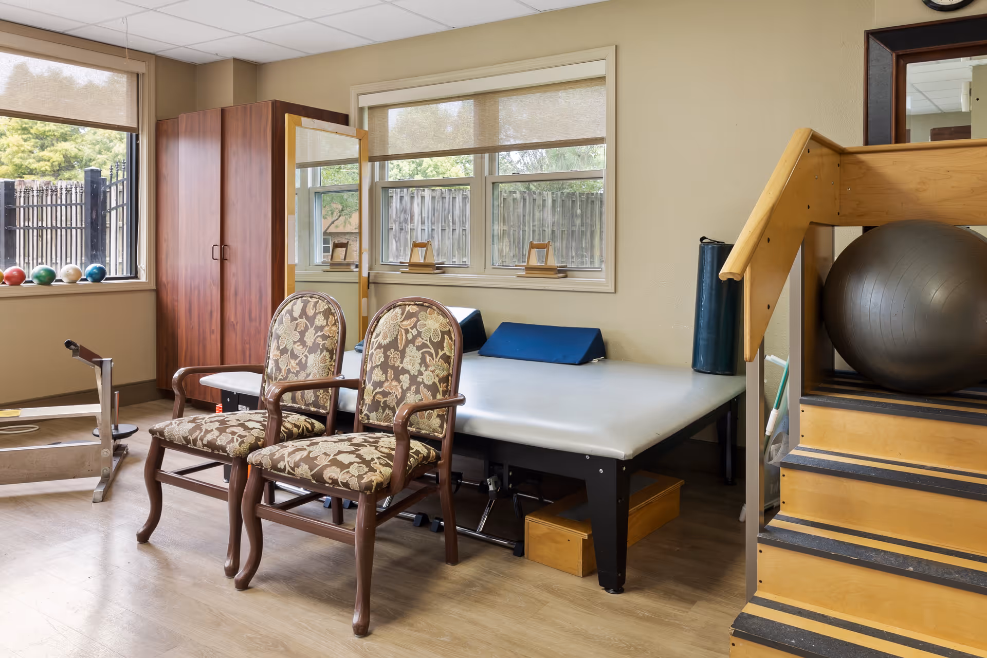 A therapy or rehabilitation room with two floral upholstered chairs, a padded therapy table, a large exercise ball on a wooden staircase, and various exercise equipment near windows with beige roller shades.