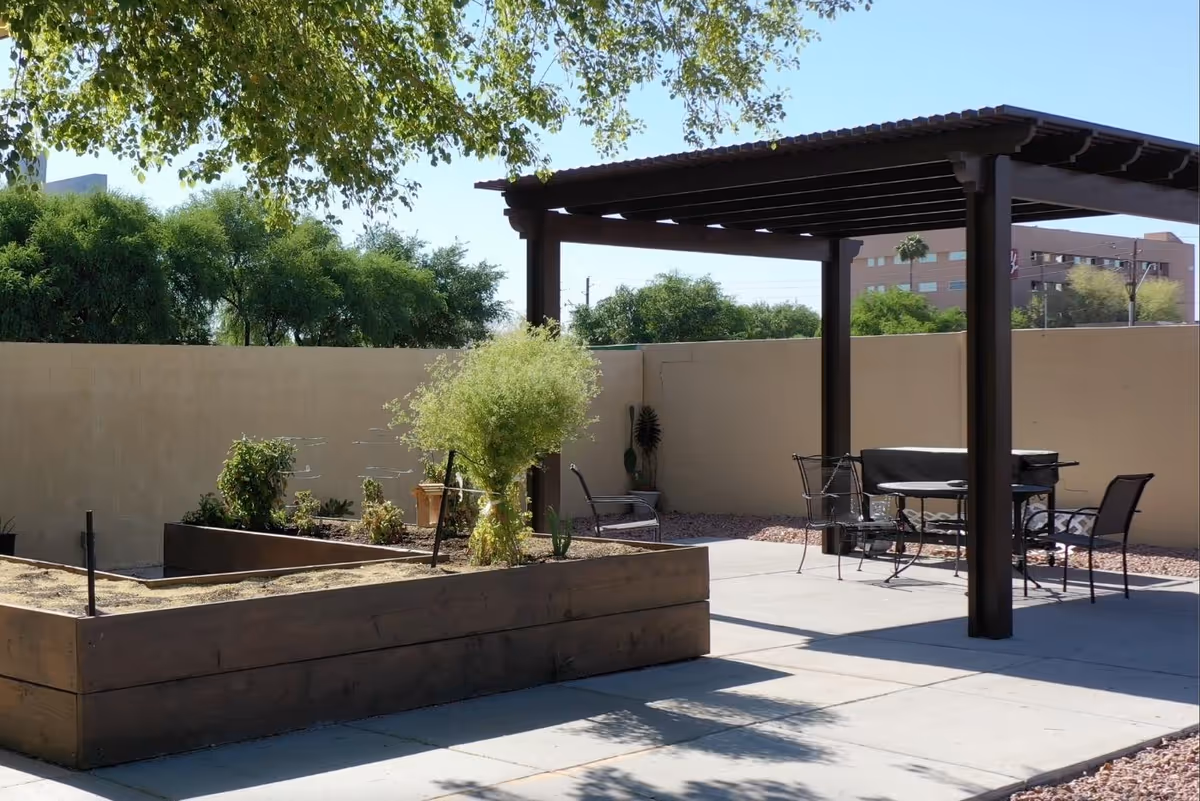 A sunlit outdoor patio with a wooden pergola, metal table and chairs, and raised planter beds against a beige wall.
