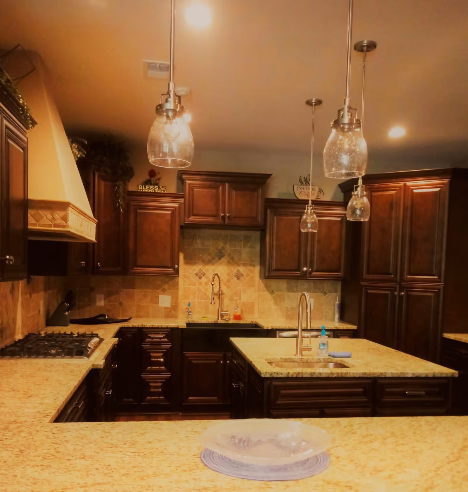 Interior view of a kitchen with dark wooden cabinets, beige granite countertops, a stove, two sinks, and three hanging pendant lights with glass shades. The backsplash features beige tiles with decorative accents.