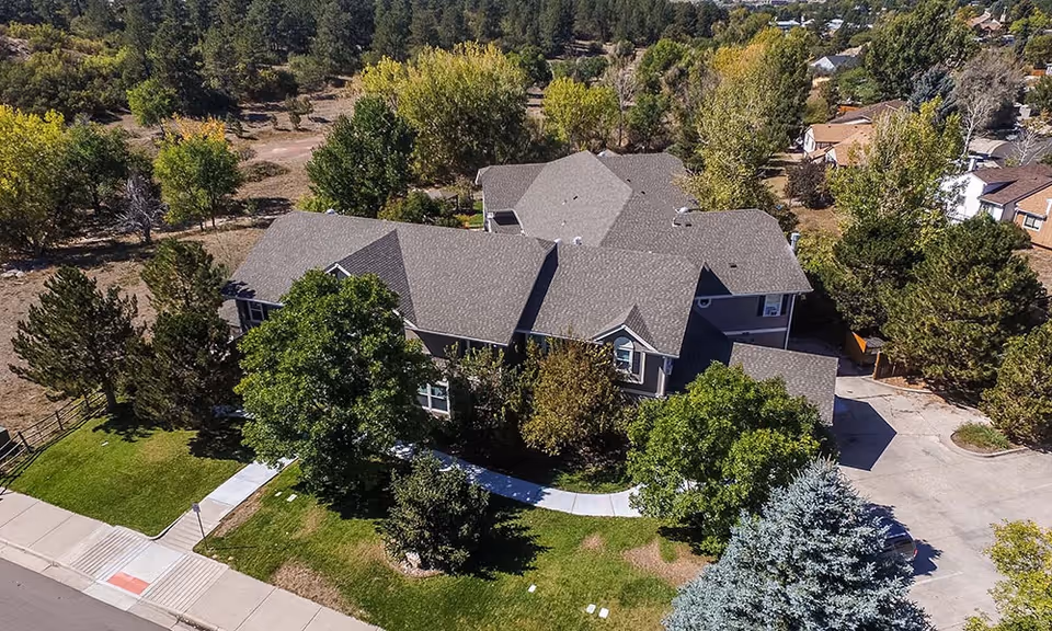 Aerial view of a multi-wing assisted living building with gray roofs surrounded by trees, lawns, sidewalks, and a driveway.