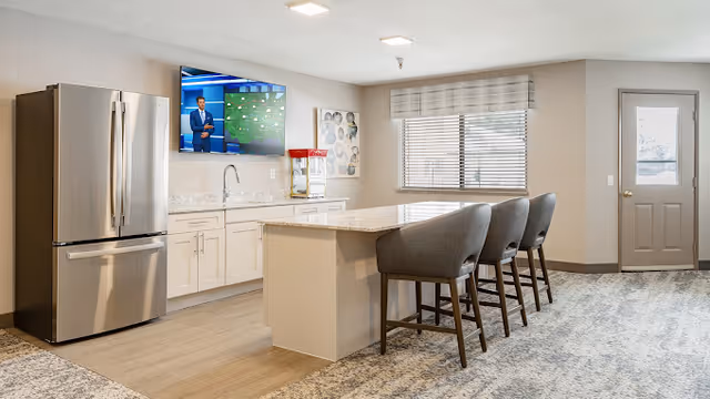 Modern kitchen area with a large island featuring a marble countertop and three gray upholstered bar stools. Stainless steel refrigerator on the left, white cabinetry, and a wall-mounted flat-screen TV displaying a weather forecast. A window with blinds and a door are visible on the right side of the room.