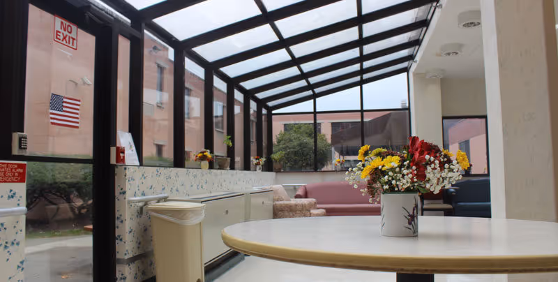 Glass-enclosed seating area with a round table topped by a vase of flowers, sofas, and windows overlooking an exterior courtyard.