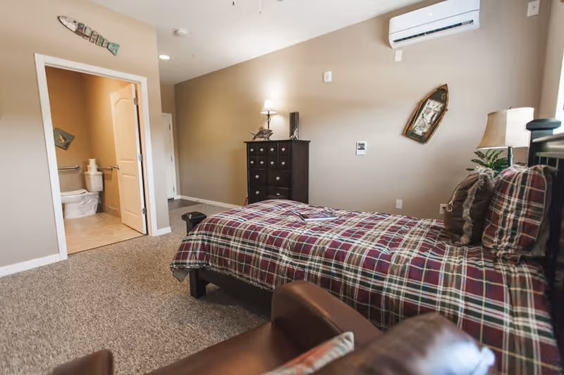 A cozy bedroom with a single bed covered in a red and green plaid comforter. There is a brown leather chair in the foreground, a dark wooden dresser with a lamp and decorative items against the wall, and a wall-mounted air conditioning unit above. The room has beige walls and carpeted floor. An open door reveals a bathroom with a toilet and towel rack.