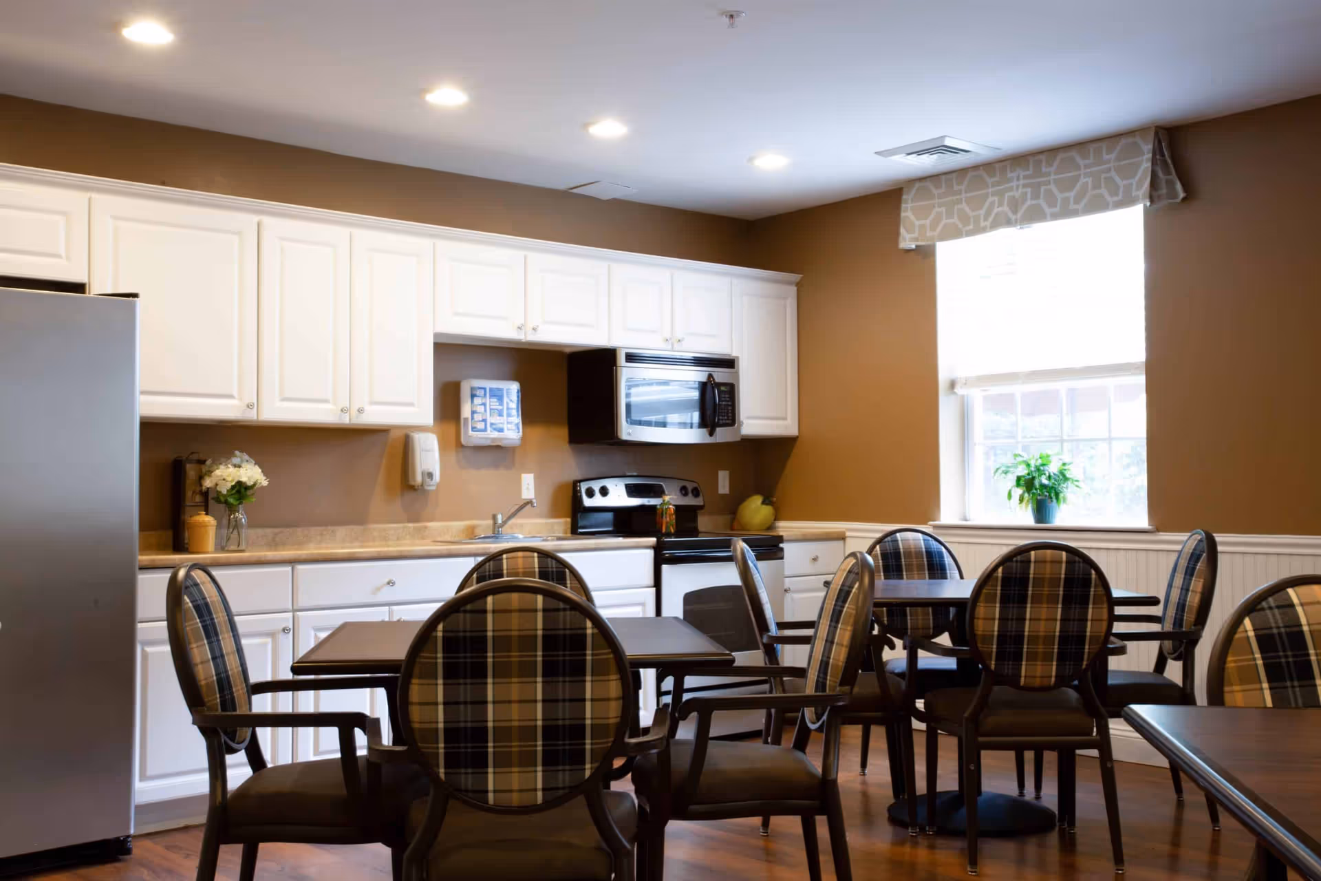 A kitchen and dining area with white cabinets, a stainless steel refrigerator, an oven with a microwave above it, and a sink. There are several tables with plaid upholstered chairs arranged around them. A window with a patterned valance and a potted plant on the windowsill lets in natural light.