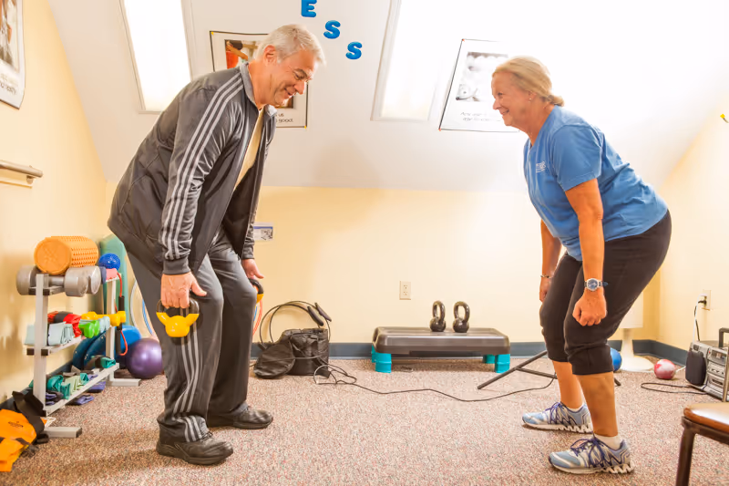 An elderly man and woman exercising together in a small fitness room. The man is holding yellow dumbbells and both are bending forward slightly, smiling. The room has exercise equipment including kettlebells, a step platform, and various small weights on shelves along the wall.
