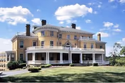 Large, historic-style yellow building with white trim and multiple chimneys under a blue sky with scattered clouds. The building has a wraparound porch with white columns and is surrounded by a well-maintained lawn and small bushes.