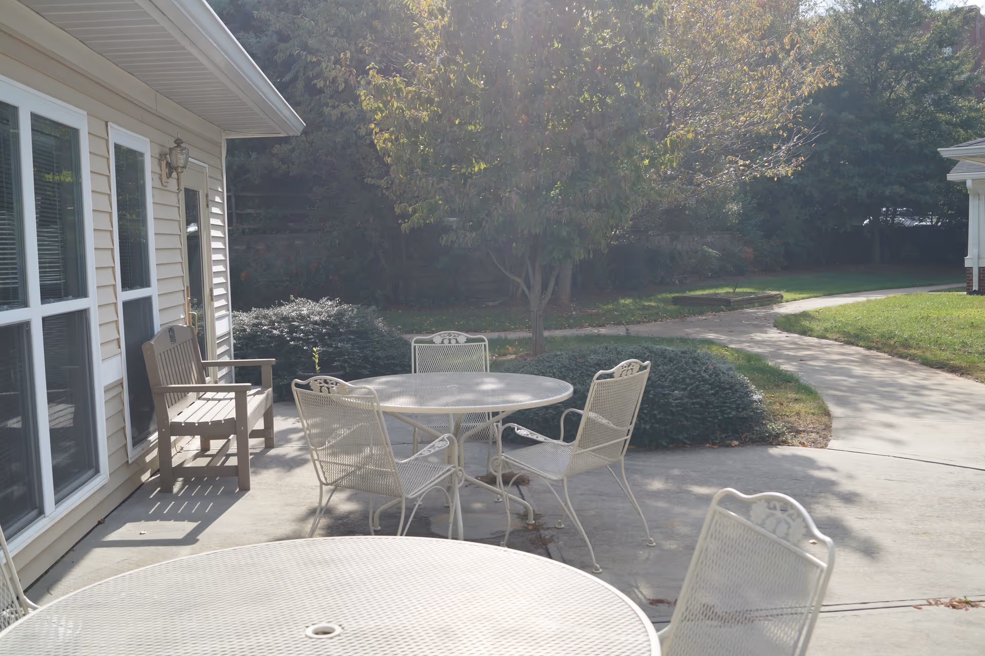 Outdoor patio area with white metal tables and chairs on a concrete surface next to a beige building with windows and a door. There is a wooden bench near the door and a tree with green foliage in the background along with bushes and a curved concrete pathway.
