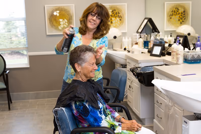 A senior woman sitting in a salon chair smiling while a hairdresser sprays her hair with hairspray in a well-lit salon area with hair care products and mirrors in the background.