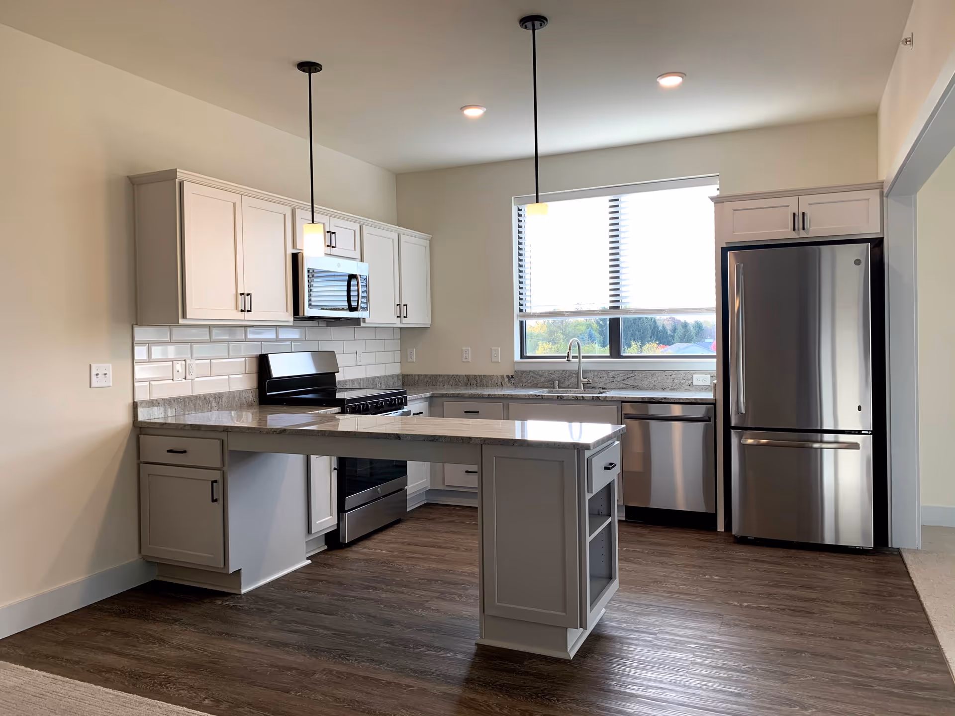 Modern kitchen with white cabinets, stainless steel appliances including a refrigerator, microwave, stove, and dishwasher. There is a kitchen island with a granite countertop, two pendant lights hanging from the ceiling, and a window with blinds letting in natural light.