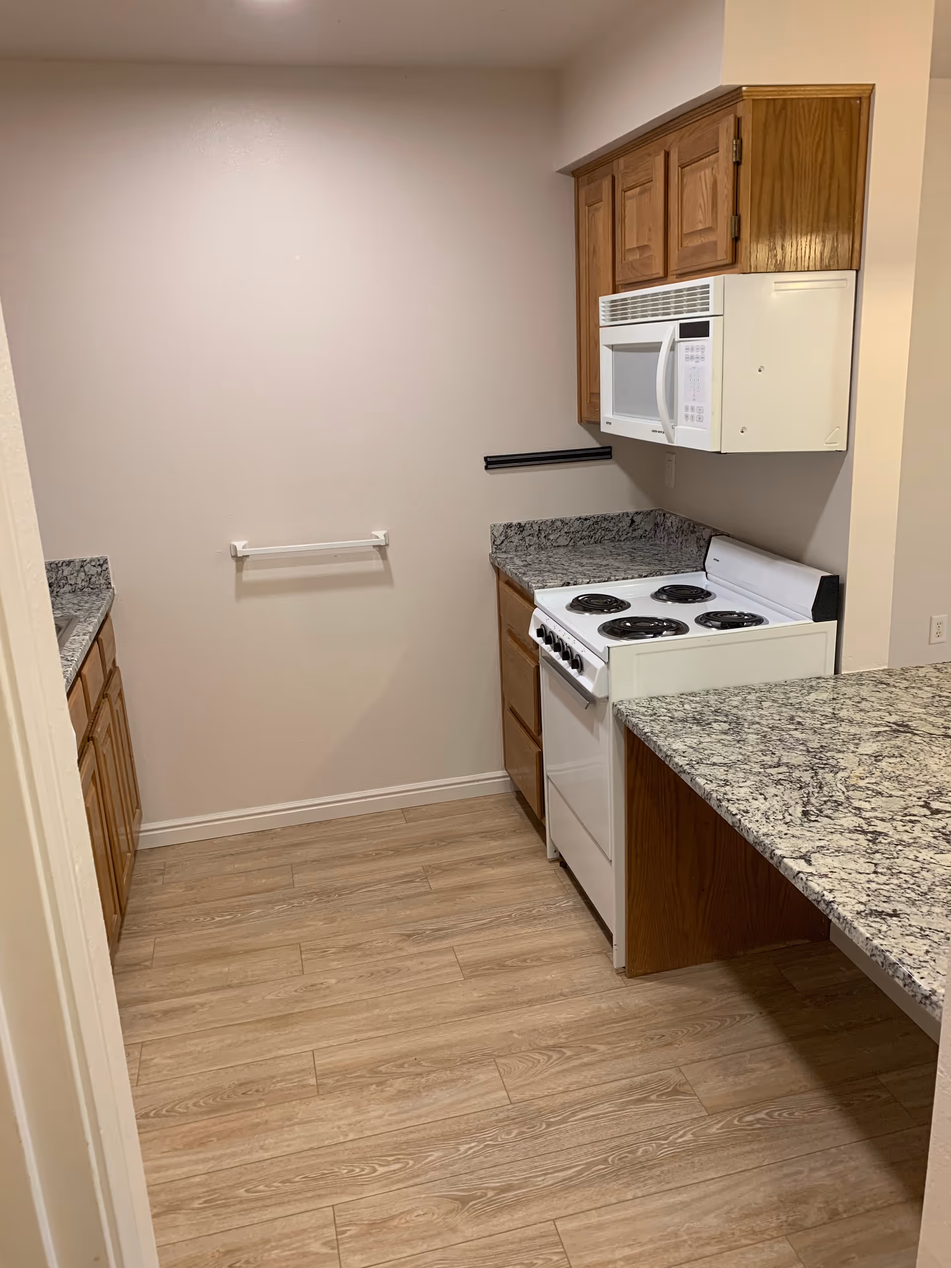 A small kitchen with wooden cabinets, granite countertops, a white electric stove with four burners, and a white microwave mounted above the stove. The floor is light wood, and the walls are painted a light beige color.