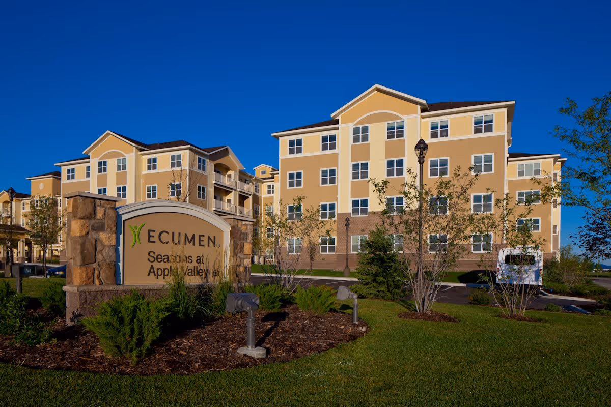 Exterior view of Ecumen Seasons at Apple Valley, a multi-story senior living facility with beige and yellow walls, multiple windows, and landscaped greenery in front including small trees and bushes under a clear blue sky.