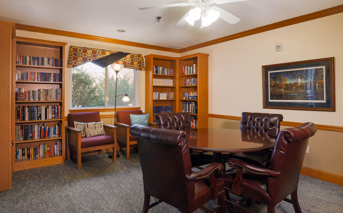 Cozy library-style common room with bookshelves, two upholstered chairs by a window, and a round wooden table surrounded by leather chairs.