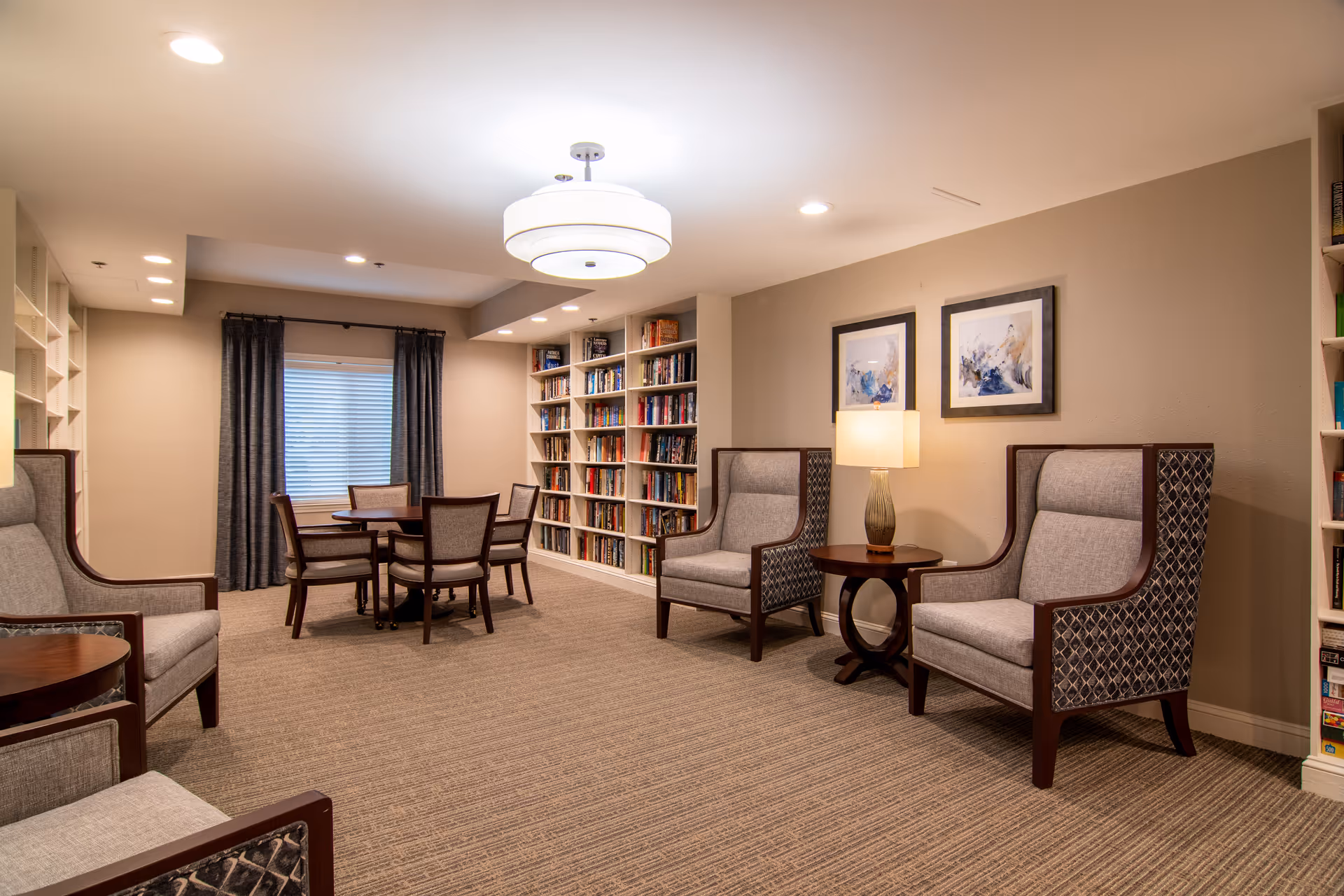 A cozy interior room with beige walls and carpeted floor featuring a seating area with four upholstered armchairs around a small wooden table with a lamp. In the background, there is a round wooden table with four chairs near a window with dark curtains. Built-in bookshelves filled with books line the walls.