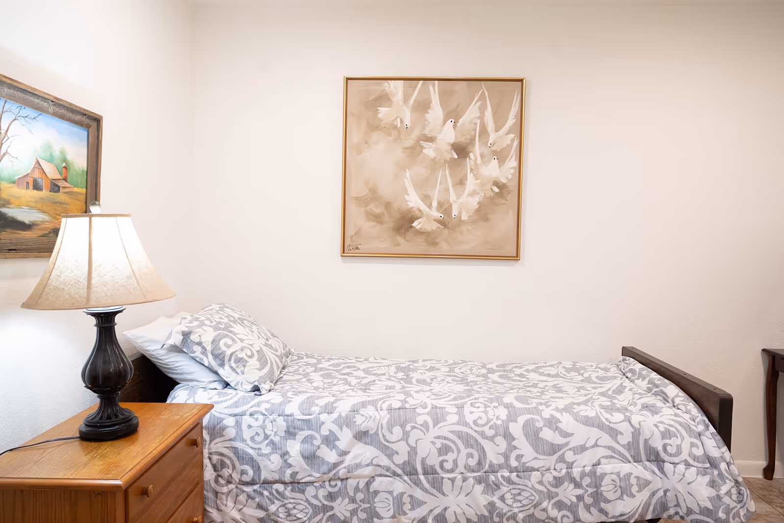 Single twin bed with patterned bedding, a wooden nightstand and lamp, and framed artwork on the wall in a simple bedroom.