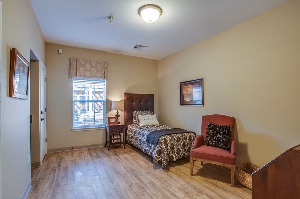 A cozy bedroom with a single bed featuring a patterned bedspread and a dark tufted headboard. Next to the bed is a wooden nightstand with a lamp and a small picture frame. A red upholstered chair with a decorative pillow is placed against the wall, and a woven basket is on the floor nearby. The room has light beige walls, a window with blinds and a valance, wooden flooring, and a framed picture hanging on the wall.