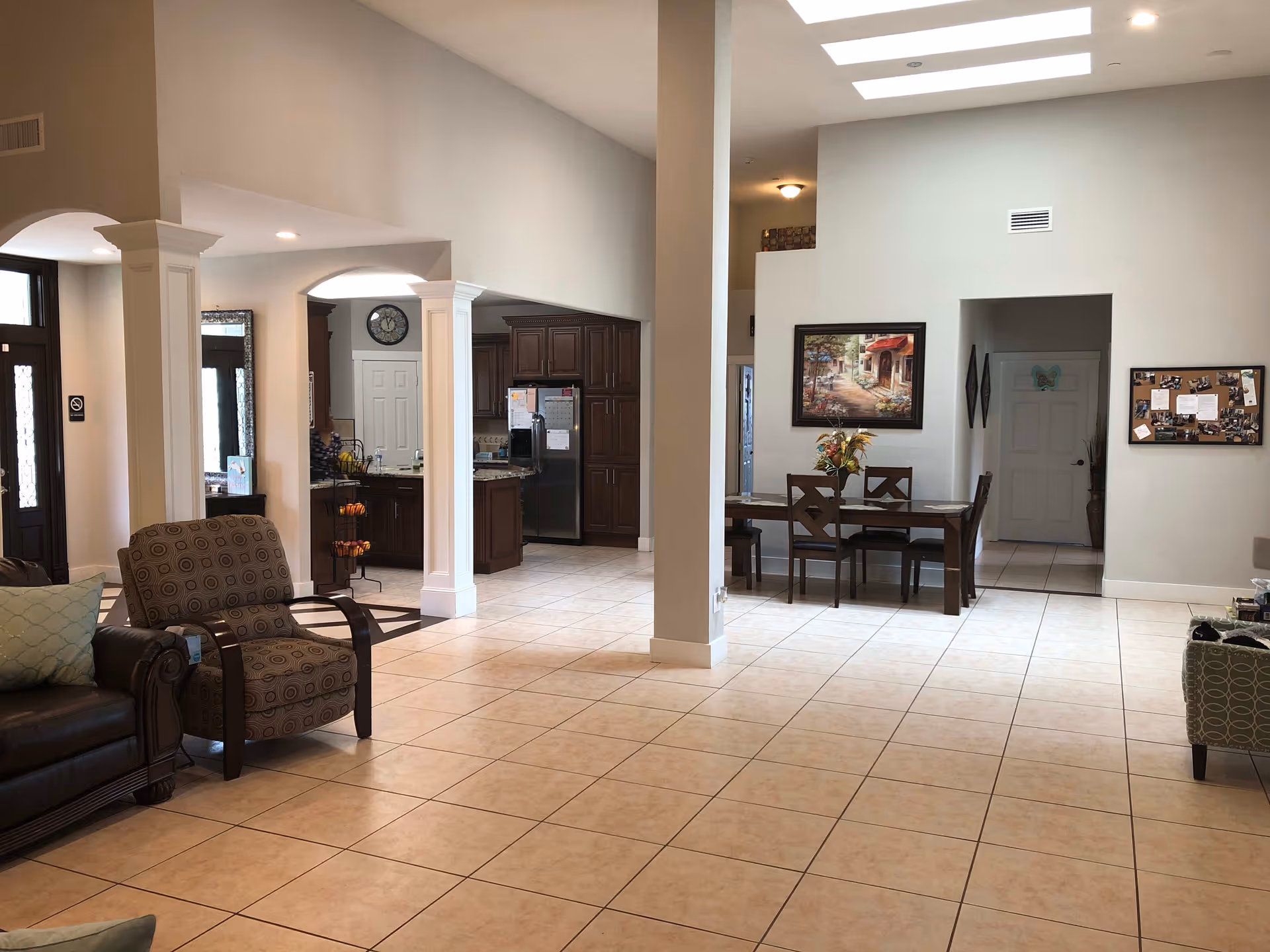 Spacious interior of a senior living facility showing a living area with a brown leather sofa and patterned armchair, a dining area with a wooden table and chairs, and a kitchen with dark wood cabinets and stainless steel refrigerator. The room has tiled floors, white walls, and skylights providing natural light.
