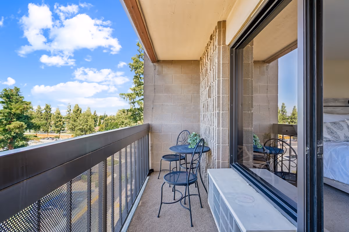 A balcony with a small round metal table and two matching chairs. The balcony has a metal railing and overlooks a view of trees and a partly cloudy sky. A sliding glass door leads to a bedroom with a bed visible inside.