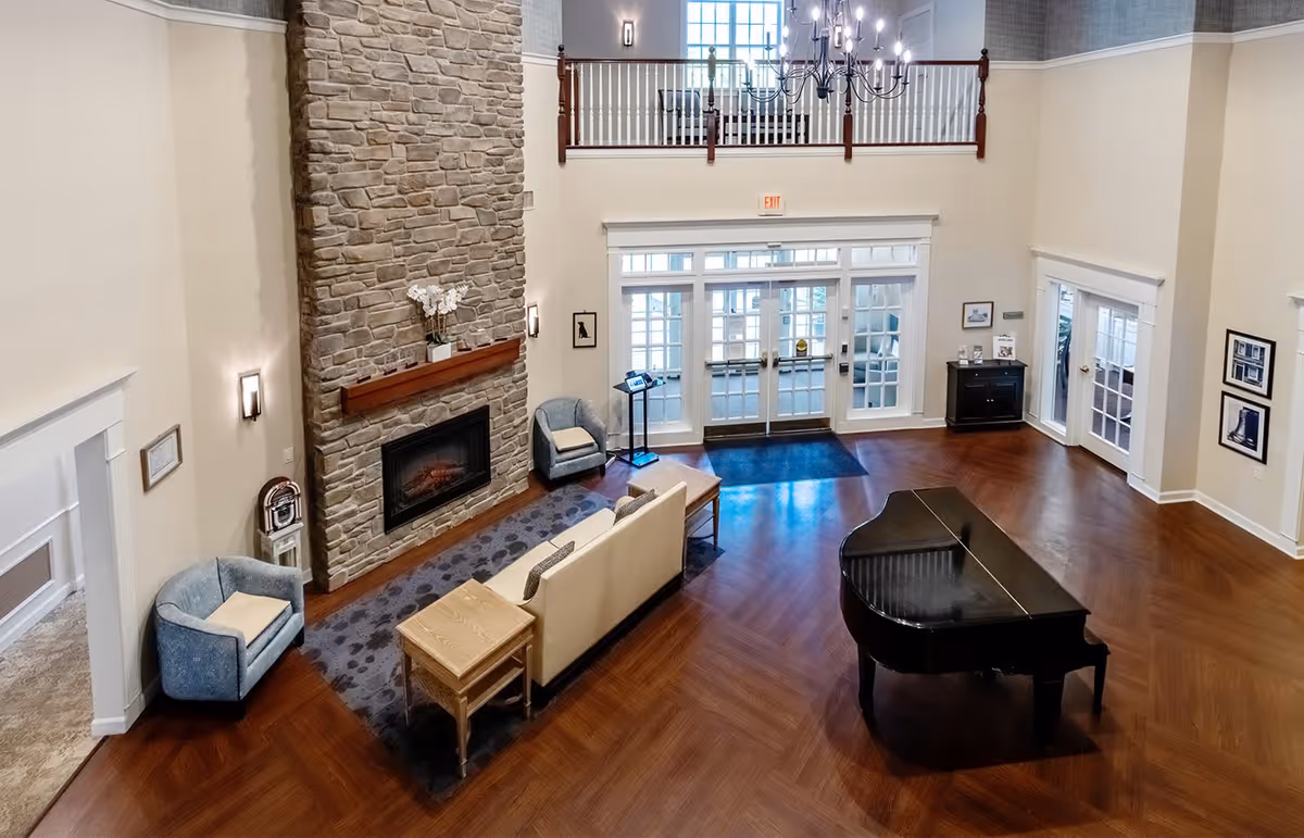 Spacious senior living facility common area with a large stone fireplace, beige sofa, two blue armchairs, a wooden side table, a black grand piano, and large windows and doors letting in natural light. The room has high ceilings with a chandelier and wooden flooring.