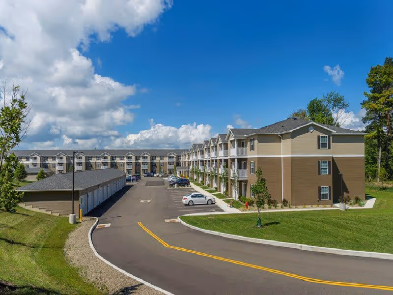 View of a multi-story residential building with balconies and a parking area in front, surrounded by green grass and trees under a partly cloudy blue sky.