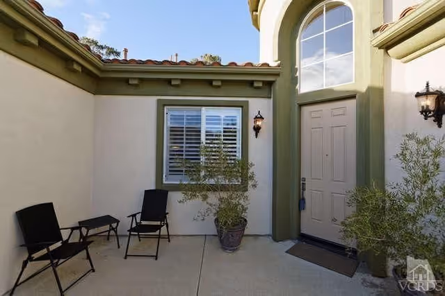Small exterior entry courtyard with two chairs and a table, potted plants, a window with shutters, and a front door with an arched transom.