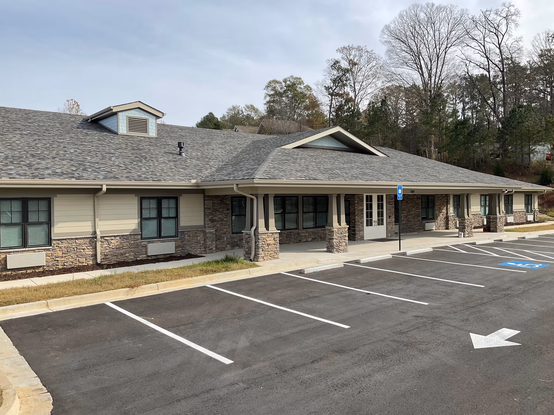 Exterior view of a single-story building with stone and beige siding, multiple windows, a covered entrance supported by stone pillars, and an empty parking lot with marked spaces including handicapped spots.