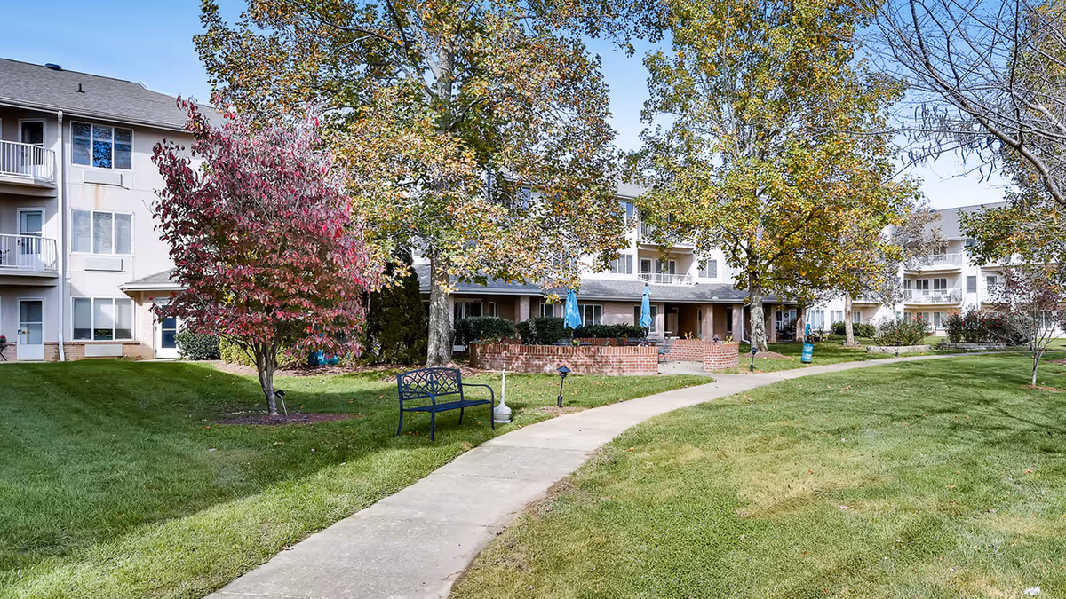 Outdoor view of a senior living community with a paved walkway curving through a grassy area, surrounded by trees with autumn foliage. There is a black metal bench along the path and multi-story residential buildings with balconies in the background.