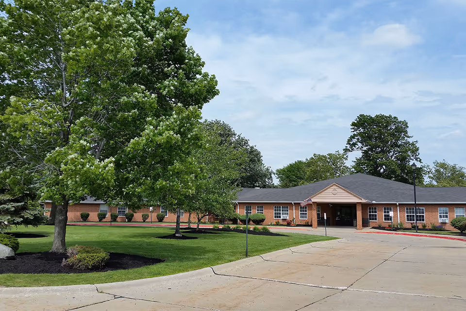 Exterior view of a single-story brick building with a dark shingled roof, surrounded by green trees and well-maintained landscaping. A paved driveway leads to the entrance, which has a covered porch and an American flag displayed near the door. The sky is partly cloudy.