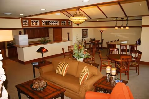 A cozy common area in a senior living facility featuring a brown sofa with striped pillows, a wooden coffee table with decorative items, an orange armchair, and several wooden dining tables and chairs. The room has warm lighting with a chandelier and pendant lights, and a small kitchen area with a refrigerator is visible in the background.