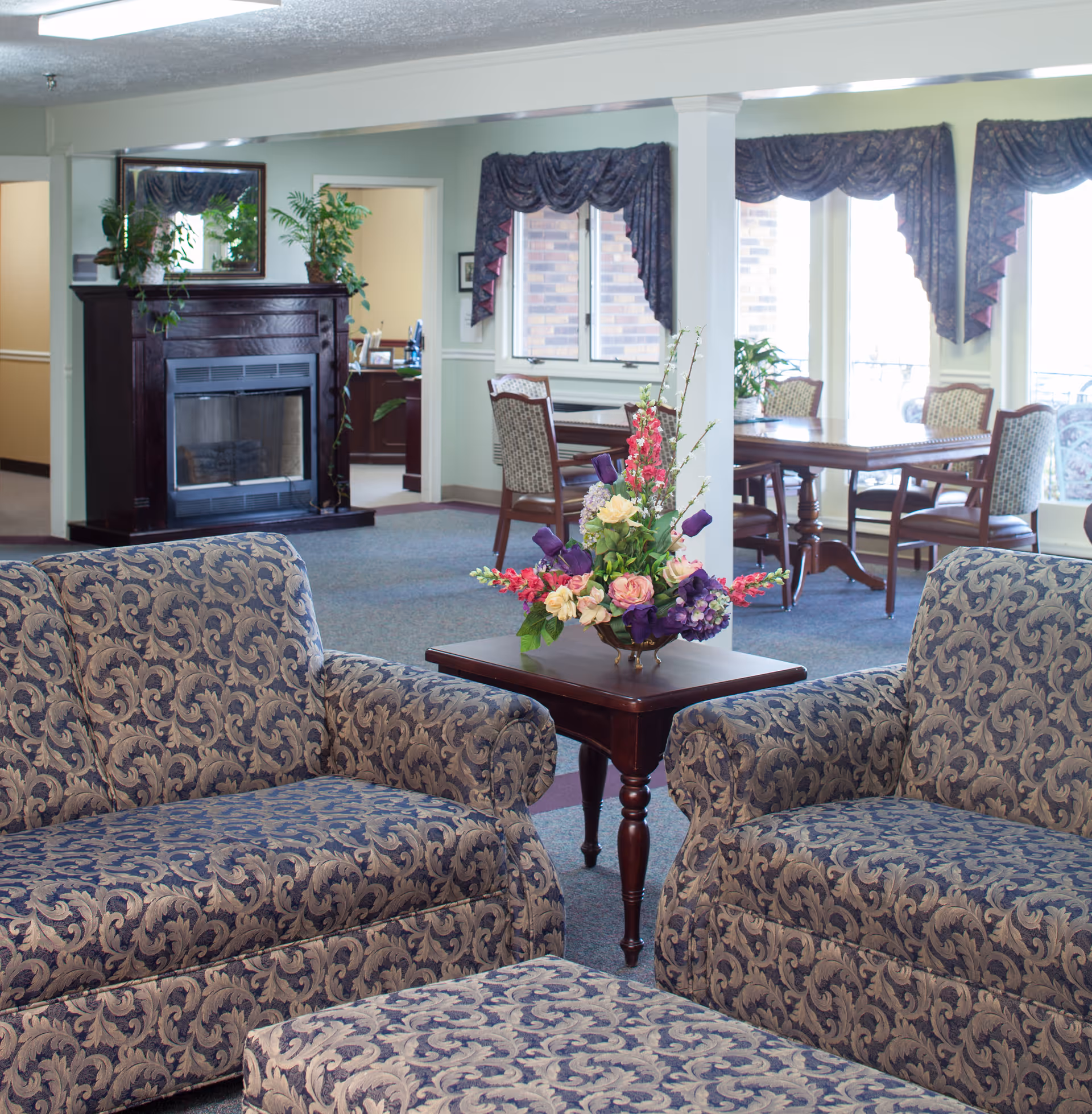 A cozy living room area with two patterned armchairs and a matching ottoman surrounding a wooden side table with a floral arrangement. In the background, there is a dark wooden fireplace with plants on top, and a dining area with tables and chairs near large windows with decorative curtains.