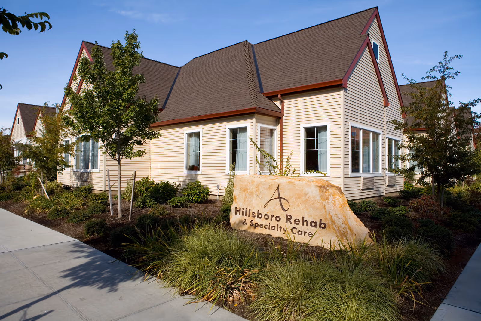 Exterior view of a beige building with brown roof and white-framed windows, surrounded by landscaped greenery and trees. A large stone sign in front reads 'Hillsboro Rehab & Specialty Care'.