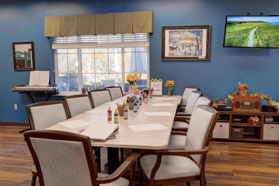A well-lit activity room with a long table surrounded by cushioned chairs. The table is set up with white paper sheets, paint bottles, and paintbrushes, suggesting an arts and crafts session. The room has blue walls, a large window with a beige valance, a keyboard on a stand, a framed painting, a flat-screen TV mounted on the wall, and a wooden cabinet decorated with autumn-themed items.