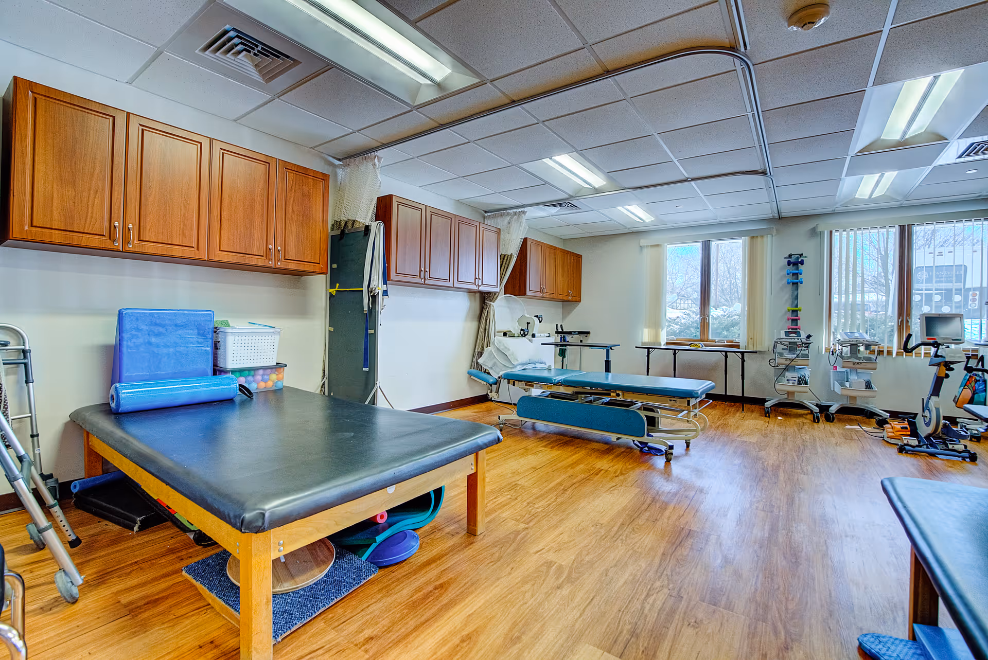 A spacious therapy or rehabilitation room with wooden flooring, two padded treatment tables, exercise equipment including a stationary bike, and wooden cabinets mounted on the walls. Large windows with vertical blinds allow natural light into the room.