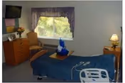 A nursing-home bedroom with a single bed covered in a blue blanket, a dresser and armchair under a wall-mounted TV, a window with purple valance, and a bedside lamp.