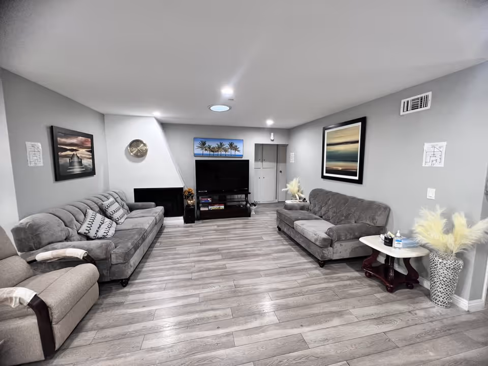 Communal living room with two gray sofas, an armchair, TV on a stand, framed artwork, and wood-look flooring.