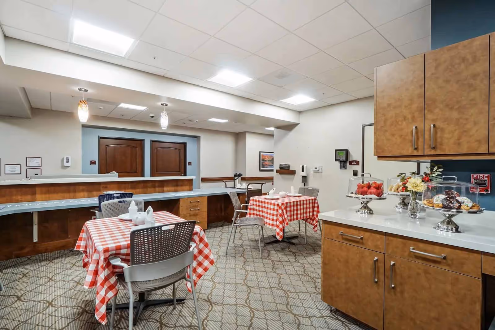 Small communal dining area with red-checkered tablecloths, chairs, a serving counter and cabinets displaying pastries and fruit.