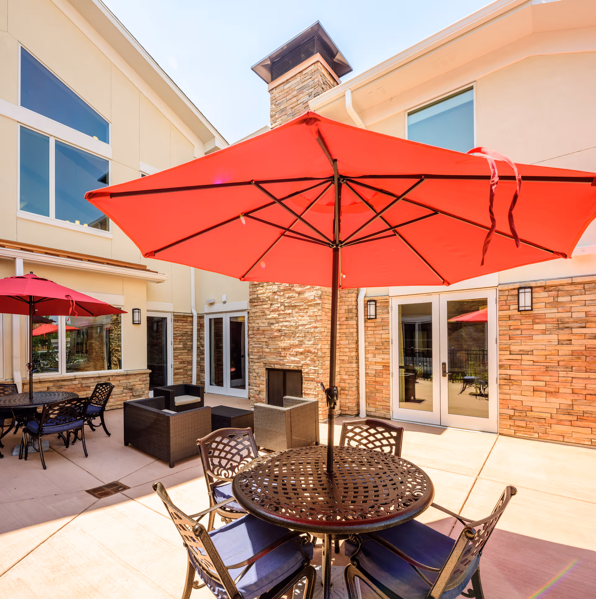 Outdoor patio area at AltaVita Assisted Living featuring round metal tables with blue cushioned chairs and large red umbrellas providing shade. The patio is surrounded by a beige building with large windows and a stone fireplace.