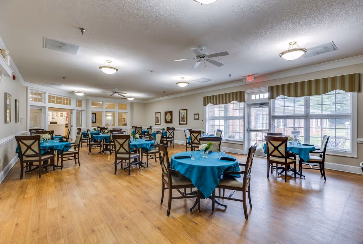 A bright dining room with multiple round tables covered with teal tablecloths, each set with chairs and small floral centerpieces. Large windows with striped valances let in natural light, and ceiling fans and light fixtures illuminate the space. The room has wooden flooring and light-colored walls with framed artwork.