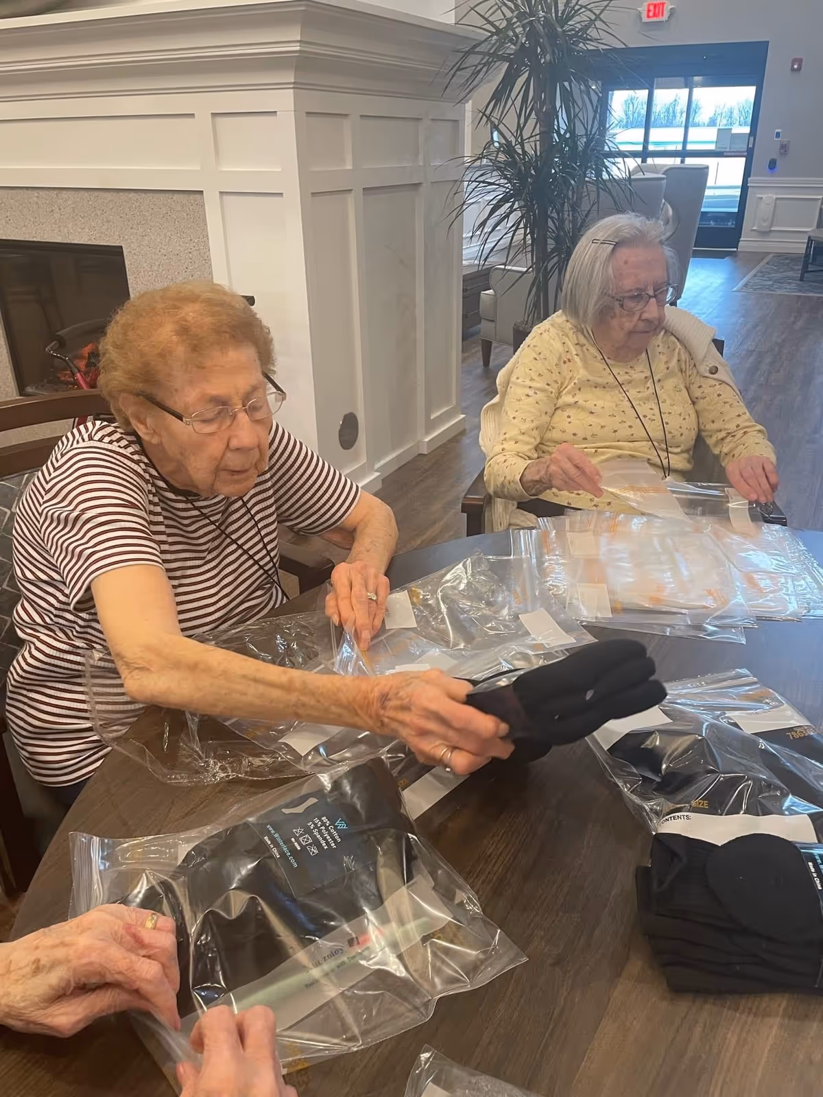 Two elderly women sitting at a wooden table inside a community room, unpacking black gloves from clear plastic bags. The room has a fireplace, large plant, and a glass door leading outside.